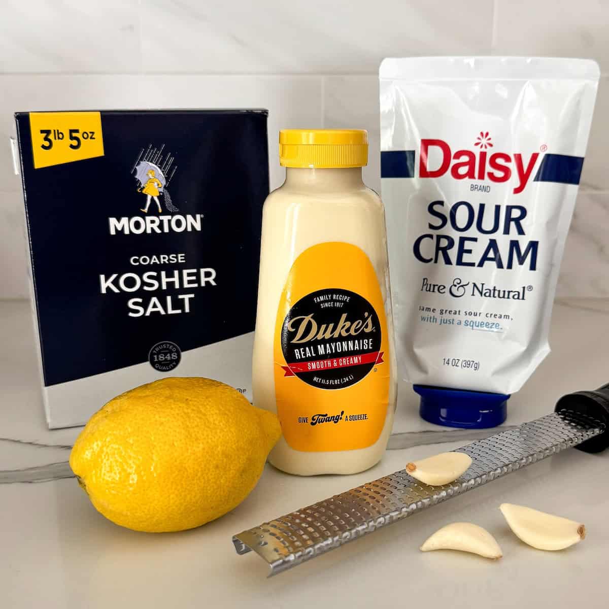 Creamy lemon aioli recipe with a bright, tangy twist! The secret is sour cream. Whisk it up in 5 minutes and keep it in the fridge for 2 weeks. Ingredients for lemon aioli recipe arranged on a countertop: a box of Morton kosher salt, a bottle of Duke’s mayonnaise, a pouch of Daisy sour cream, a lemon, a metal zester, and three garlic cloves.