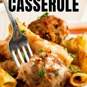 A fork holds a cheesy meatball over a serving of cheesy pasta and meatball casserole on a plate, garnished with herbs.