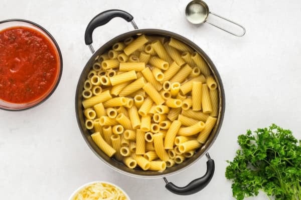 With seared meatballs, lots of cheese, and perfectly cooked saucy pasta (that's never mushy), this is the best Meatball Casserole recipe! A pot filled with cooked rigatoni pasta is on a white surface, surrounded by a bowl of tomato sauce, shredded cheese, a metal measuring cup, and a bunch of fresh parsley.