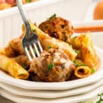 A close-up of a plate of baked ziti pasta with meatballs, melted cheese, and garnished with parsley. A hand holds a fork, about to take a bite.