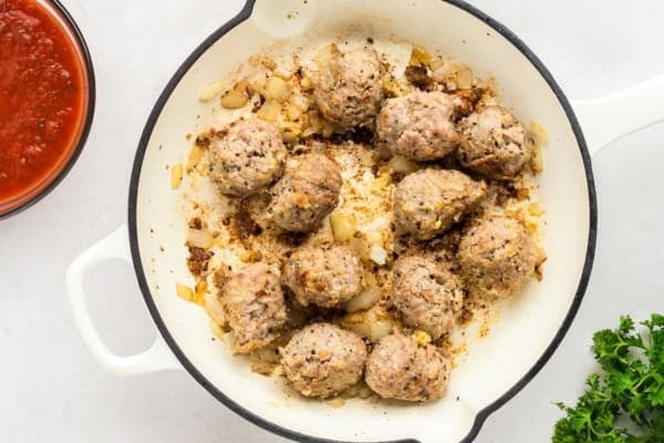 With seared meatballs, lots of cheese, and perfectly cooked saucy pasta (that's never mushy), this is the best Meatball Casserole recipe! A white skillet containing browned meatballs with sautéed onions sits on a white surface. A bowl of marinara sauce and a bunch of fresh parsley are placed nearby.