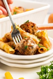 A close-up of a plate of baked ziti pasta with meatballs, melted cheese, and garnished with parsley. A hand holds a fork, about to take a bite.