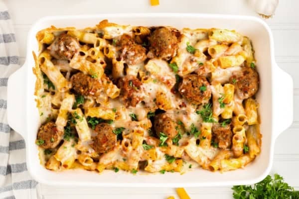 With seared meatballs, lots of cheese, and perfectly cooked saucy pasta (that's never mushy), this is the best Meatball Casserole recipe! A baked meatball casserole in a white casserole dish, featuring ziti pasta, meatballs, melted cheese, and sprinkled with chopped parsley. The dish sits on a white surface with a striped cloth nearby.