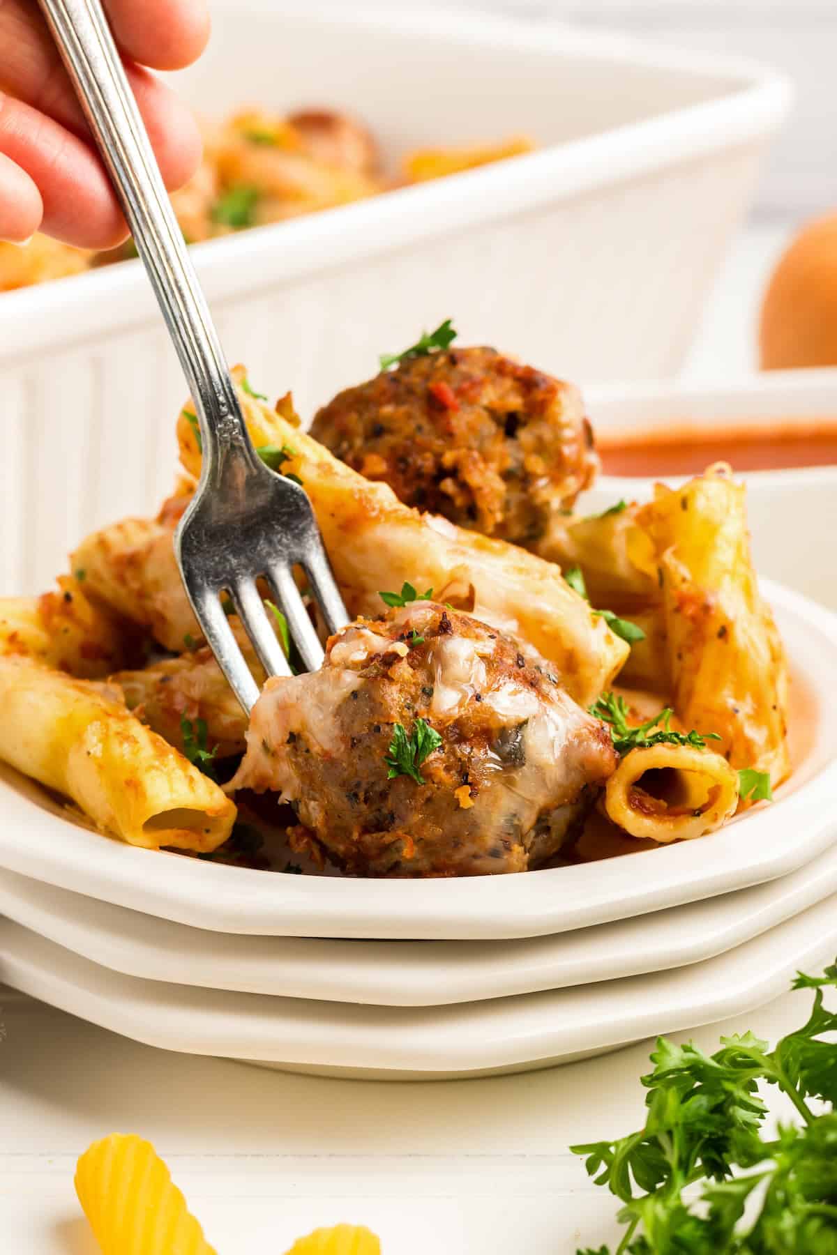 With seared meatballs, lots of cheese, and perfectly cooked saucy pasta (that's never mushy), this is the best Meatball Casserole recipe! A close-up of a plate of baked ziti with meatballs, melted cheese, and garnished with parsley. A hand holds a fork, about to take a bite.