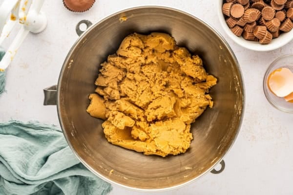 These giant bakery-style Reese's Peanut Butter Cookies are loaded with mini cups inside and a full size Reese's cup melted on top! A mixing bowl filled with peanut butter cookie dough sits on a light countertop, surrounded by a blue cloth, a cracked egg in a dish, a bowl of mini peanut butter cups, and a mixer attachment.