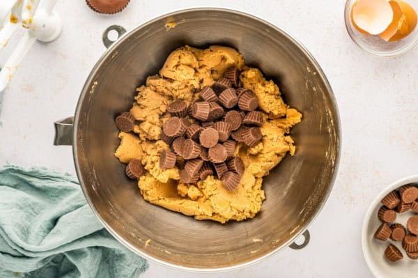 These giant bakery-style Reese's Peanut Butter Cookies are loaded with mini cups inside and a full size Reese's cup melted on top! A metal mixing bowl filled with peanut butter cookie dough and mini peanut butter cups sits on a light countertop.
