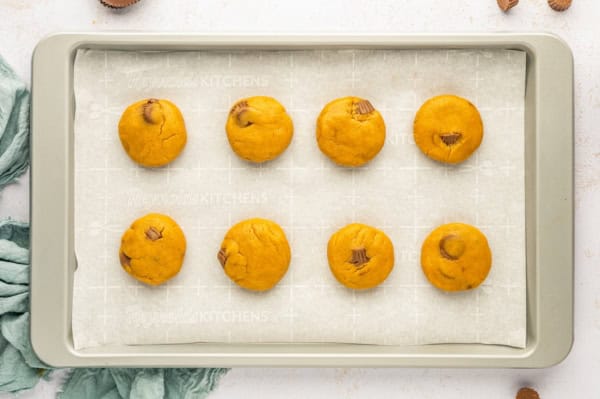 These giant bakery-style Reese's Peanut Butter Cookies are loaded with mini cups inside and a full size Reese's cup melted on top! A baking sheet lined with parchment paper holds eight flattened balls of peanut butter cookie dough stuffed with mini Reese's cups. The cookies are evenly spaced in two rows of four.