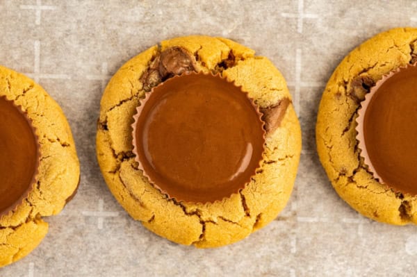 These giant bakery-style Reese's Peanut Butter Cookies are loaded with mini cups inside and a full size Reese's cup melted on top! A close-up of three Reeses peanut butter cookies, each topped with a round full-size chocolate peanut butter cup, on a sheet of parchment paper.
