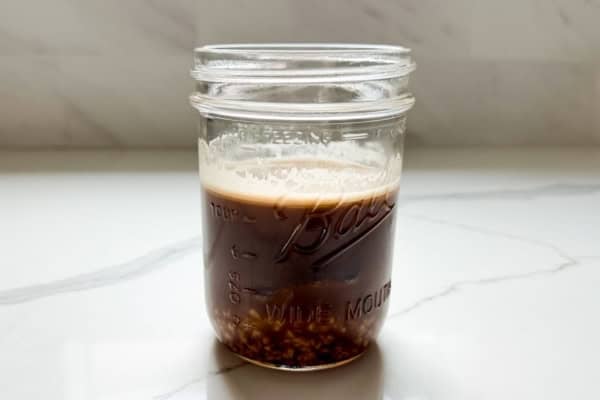 Sticky Toffee Date Coffee Syrup is made with 3 ingredients and NO cooking. Soak dates and toffee bits in hot espresso, blend, and enjoy! A glass Mason jar containing a espresso with toffee bits and dates settled at the bottom, placed on a white marble countertop.