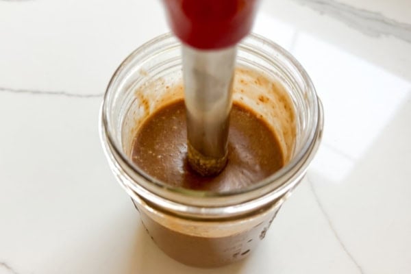 Sticky Toffee Date Coffee Syrup is made with 3 ingredients and NO cooking. Soak dates and toffee bits in hot espresso, blend, and enjoy! A handheld immersion blender is mixing a thick coffee date syrup in a glass jar on a white countertop.