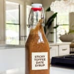 A glass bottle labeled Sticky Toffee Date Syrup filled with thick brown homemade coffee syrup sits on a white kitchen counter, with a bright modern kitchen and large windows in the background.