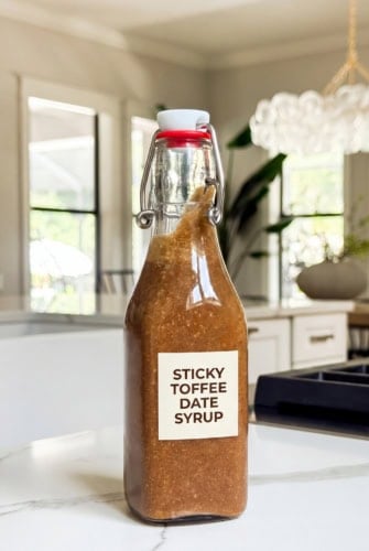 A glass bottle labeled Sticky Toffee Date Syrup filled with thick brown homemade coffee syrup sits on a white kitchen counter, with a bright modern kitchen and large windows in the background.