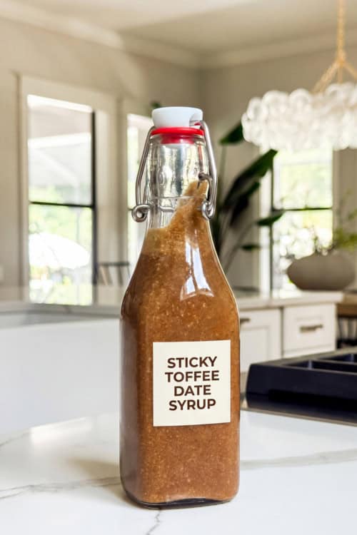 A glass bottle labeled Sticky Toffee Date Syrup filled with thick brown homemade coffee syrup sits on a white kitchen counter, with a bright modern kitchen and large windows in the background.