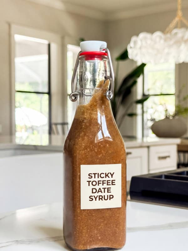 A glass bottle labeled Sticky Toffee Date Syrup filled with thick brown homemade coffee syrup sits on a white kitchen counter, with a bright modern kitchen and large windows in the background.