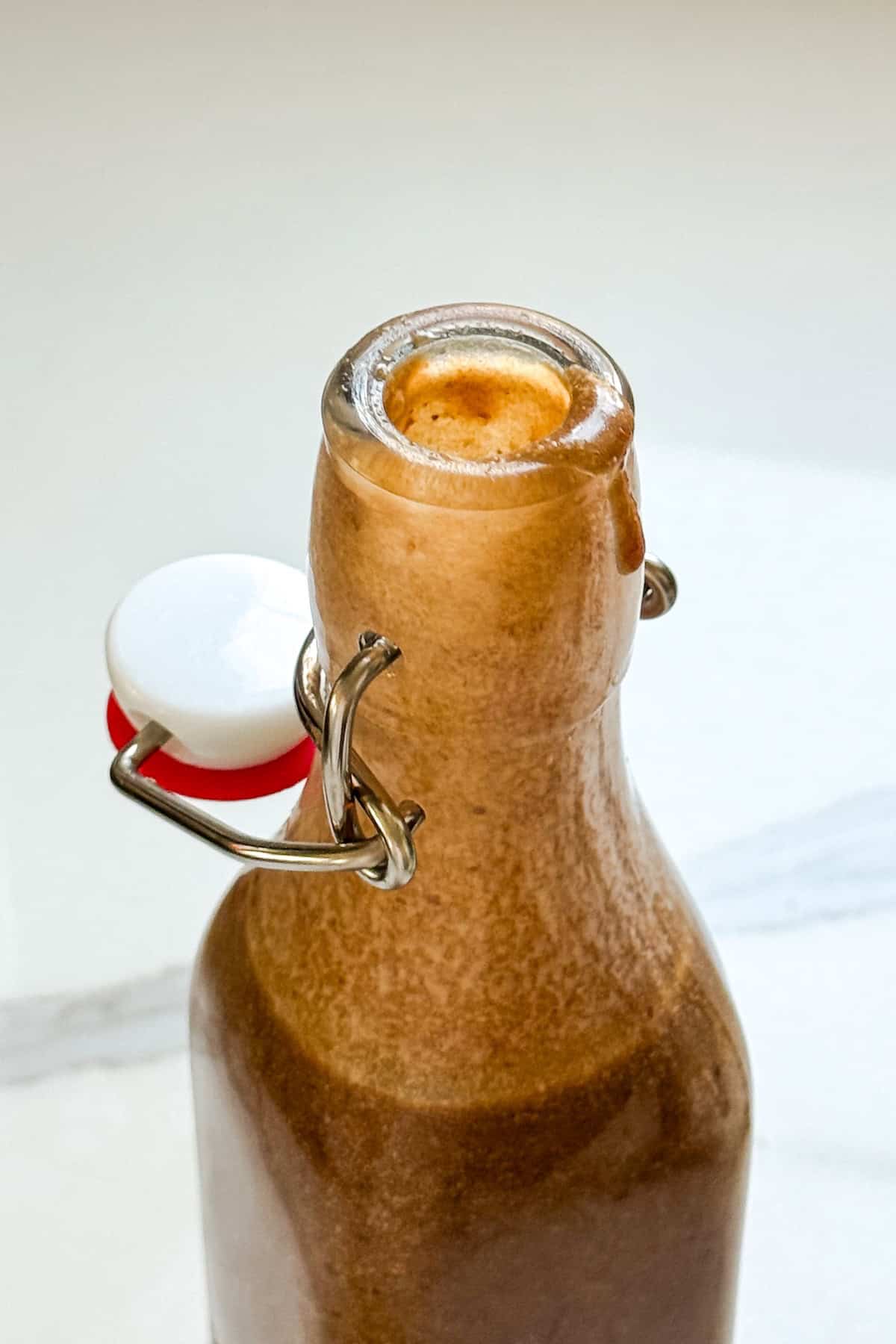 Sticky Toffee Date Coffee Syrup is made with 3 ingredients and NO cooking. Soak dates and toffee bits in hot espresso, blend, and enjoy! A glass bottle filled with a thick, brown sticky toffee date coffee syrup, slightly overflowing at the rim, with a metal clasp and white ceramic stopper on the side. The bottle sits on a light, smooth surface.