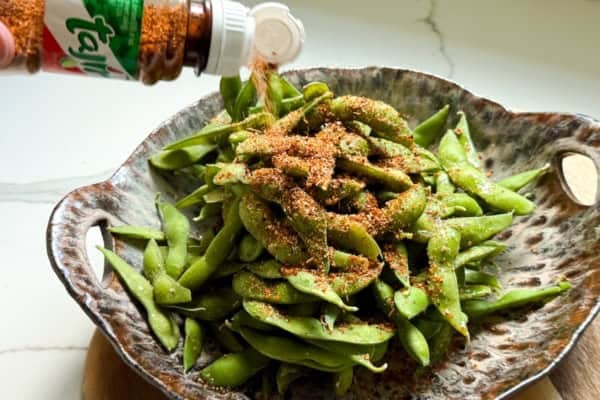 Make the BEST spicy edamame recipe at home with frozen pods, fresh lime, and Tajin. Easy, addictive, and ready in just 5 minutes! A hand sprinkles Tajín seasoning from a bottle onto a mound of steamed edamame in a decorative bowl on a wooden surface.