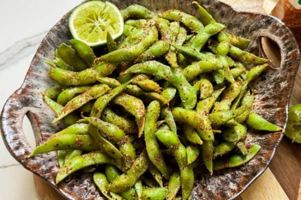 Make the BEST spicy edamame recipe at home with frozen pods, fresh lime, and Tajin. Easy, addictive, and ready in just 5 minutes! A rustic bowl filled with spicy edamame pods sprinkled with lime and spices with a halved lime on the side.