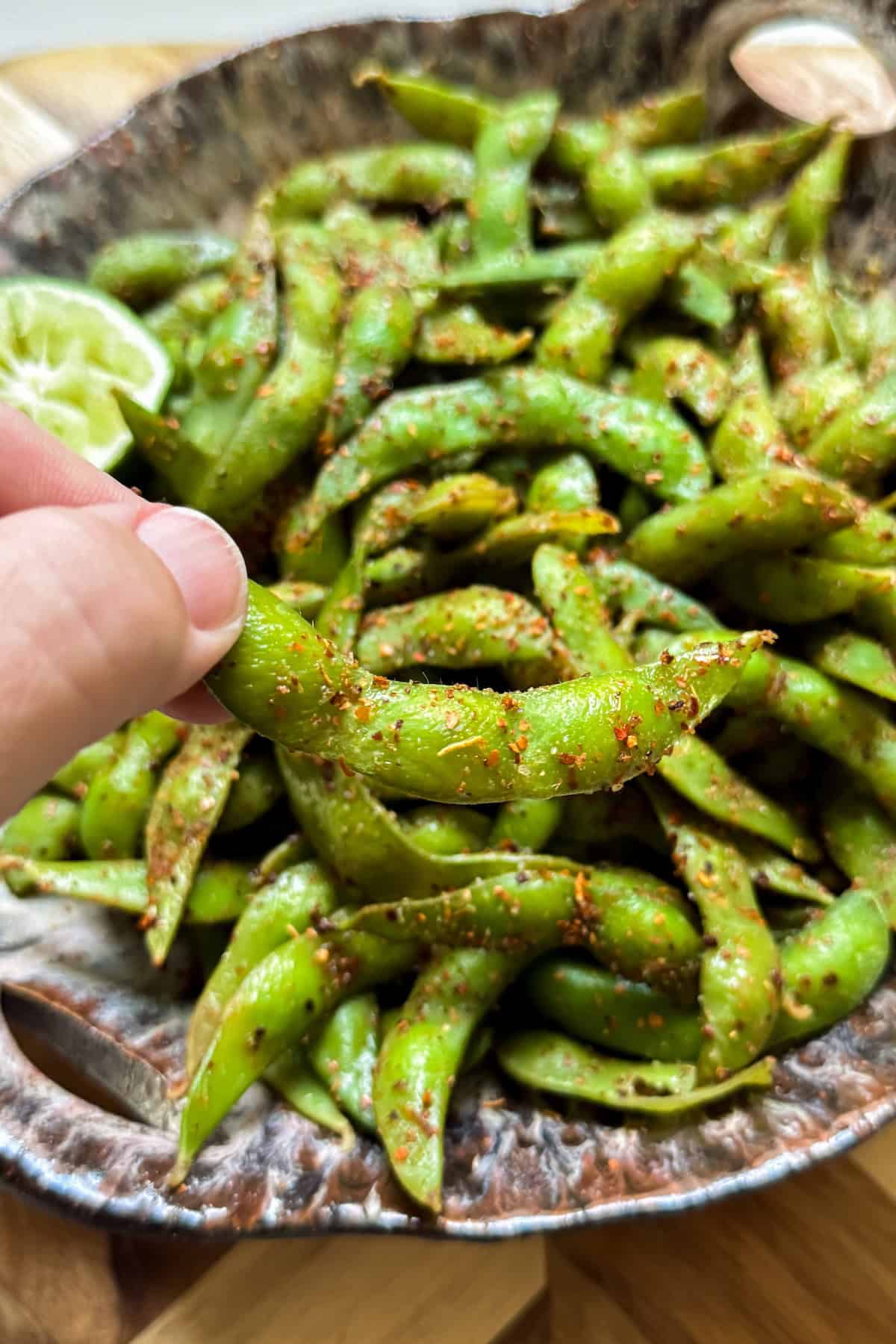 Make the BEST spicy edamame recipe at home with frozen pods, fresh lime, and Tajin. Easy, addictive, and ready in just 5 minutes! A hand holds a seasoned spicy edamame pod above a rustic bowl filled with more chili-dusted edamame. A lime wedge is visible in the bowl.