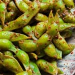 A close-up of a plate filled with bright green spicy edamame pods coated in lime juice and chili seasoning.