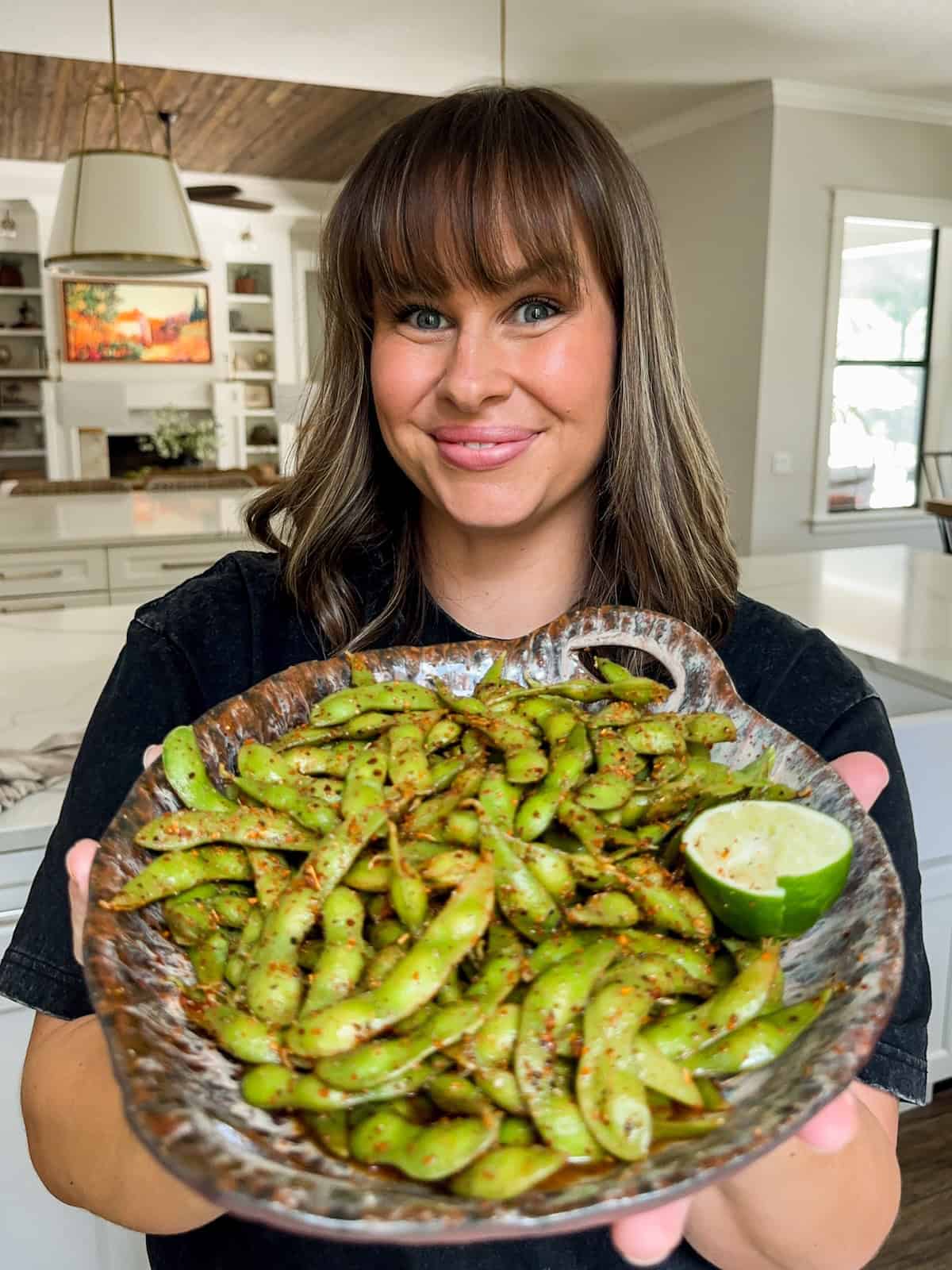 Make the BEST spicy edamame recipe at home with frozen pods, fresh lime, and Tajin. Easy, addictive, and ready in just 5 minutes! A smiling woman in a kitchen holds a rustic plate filled with spicy chili lime edamame pods with a halved lime on the side. The background shows a modern, bright kitchen with white cabinets.