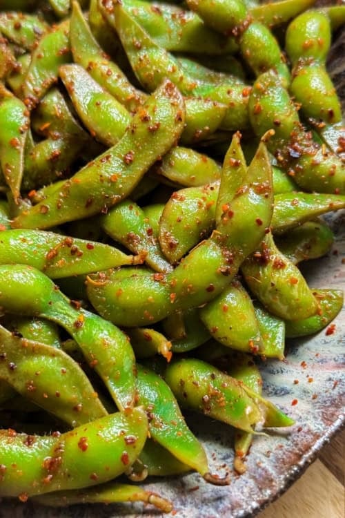A close-up of a plate filled with bright green spicy edamame pods coated in lime juice and chili seasoning.