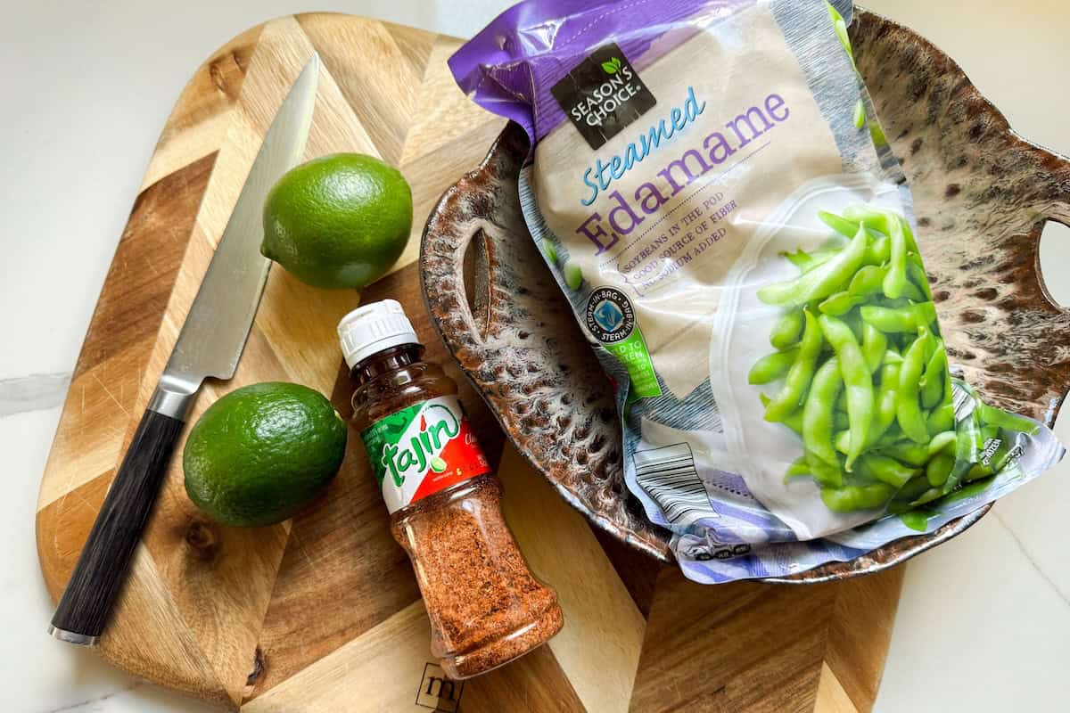 Make the BEST spicy edamame recipe at home with frozen pods, fresh lime, and Tajin. Easy, addictive, and ready in just 5 minutes! A cutting board with a knife and two limes, next to a bottle of Tajín seasoning and a bag of frozen edamame in a bowl.