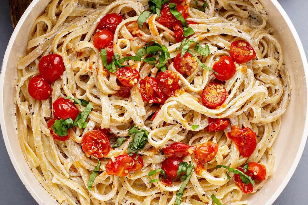 This creamy Cherry Tomato Pasta recipe is loaded with garlic roasted tomatoes and a tangy goat cheese sauce. An easy dinner ready in 30 minutes! A bowl of creamy tomato pasta topped with roasted cherry tomatoes and fresh basil, garnished with herbs.