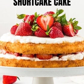 A homemade strawberry shortcake cake with two layers, whipped cream, and fresh strawberries on top, displayed on a white cake stand.