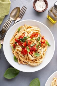A white plate of creamy cherry tomato pasta topped with fresh basil sits on a gray table beside basil leaves, a fork, spoon, a bowl of salt, and a bottle of olive oil.