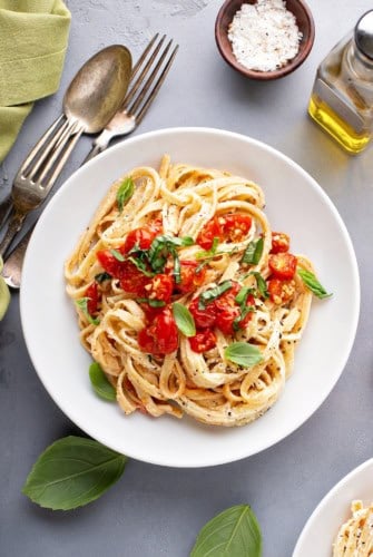 A white plate of creamy cherry tomato pasta topped with fresh basil sits on a gray table beside basil leaves, a fork, spoon, a bowl of salt, and a bottle of olive oil.