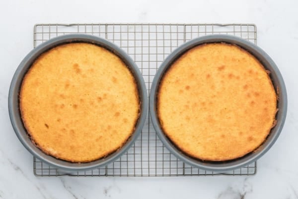 This homemade Strawberry Shortcake Cake recipe stacks a dense and rich butter cake with whipped cream and macerated strawberries in every bite. Two round, golden-brown cakes in metal pans are cooling on a wire rack placed on a white marble surface.