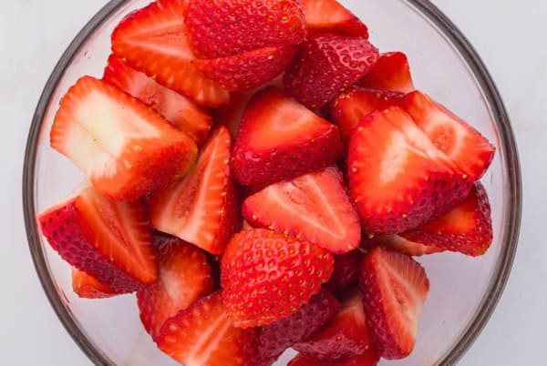 This homemade Strawberry Shortcake Cake recipe stacks a dense and rich butter cake with whipped cream and macerated strawberries in every bite. A glass bowl filled with fresh, halved and quartered strawberries on a white surface.