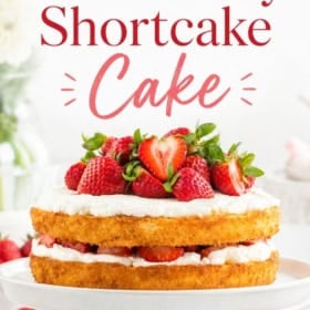 A homemade strawberry shortcake cake with two layers, cream, and fresh strawberries sits on a white cake stand.