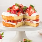 A two-layer homemade strawberry shortcake cake topped and filled with whipped cream and fresh strawberries sits on a white cake stand, with one large slice removed, revealing the inside.