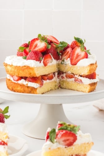 A two-layer homemade strawberry shortcake cake topped and filled with whipped cream and fresh strawberries sits on a white cake stand, with one large slice removed, revealing the inside.