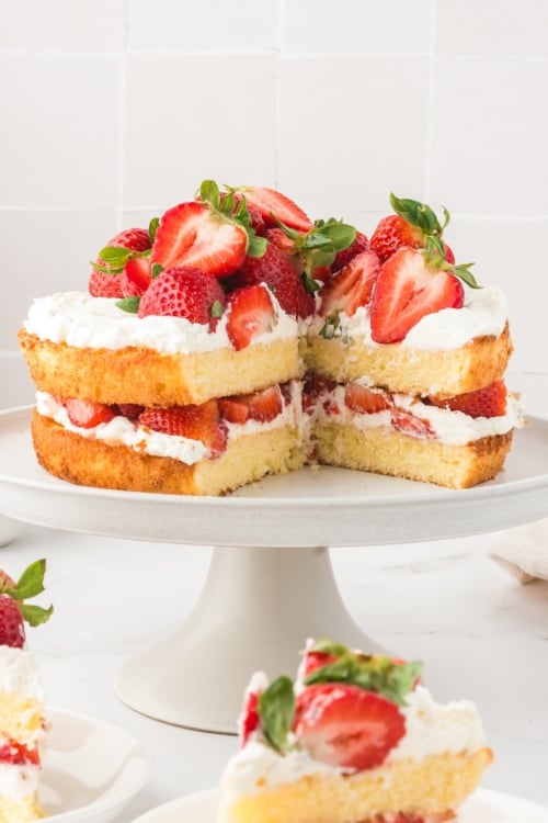 A two-layer homemade strawberry shortcake cake topped and filled with whipped cream and fresh strawberries sits on a white cake stand, with one large slice removed, revealing the inside.