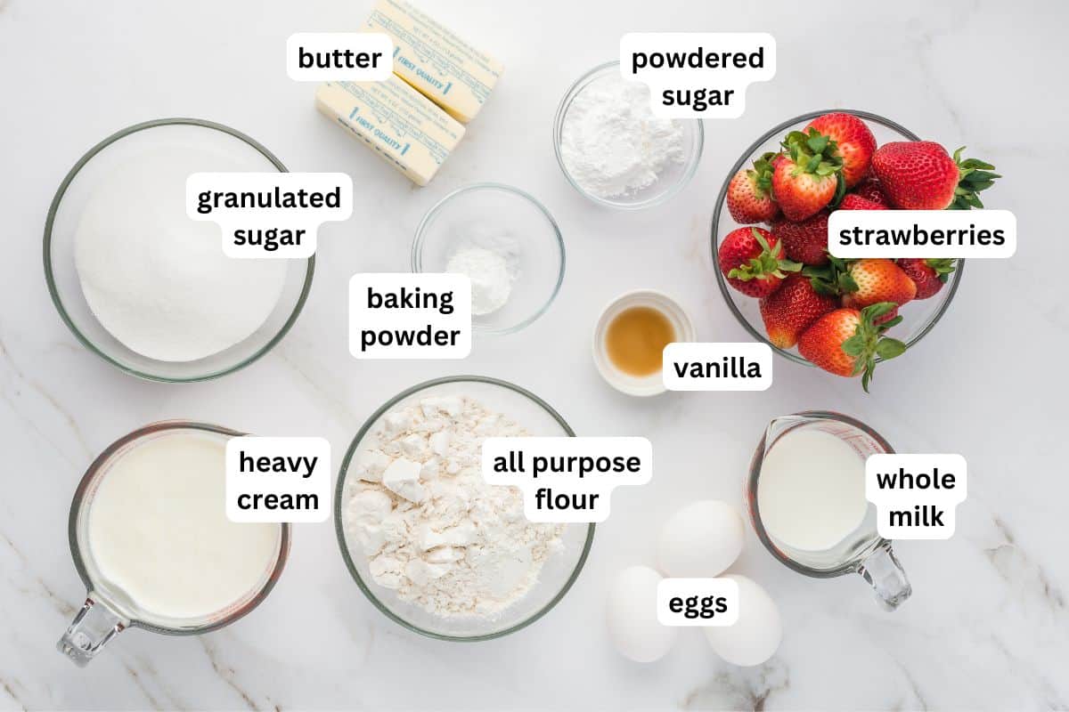 This homemade Strawberry Shortcake Cake recipe stacks a dense and rich butter cake with whipped cream and macerated strawberries in every bite. Labeled ingredients for a strawberry shortcake cake recipe arranged on a white surface: butter, powdered sugar, strawberries, granulated sugar, baking powder, vanilla, heavy cream, all purpose flour, eggs, and whole milk in bowls or cups.