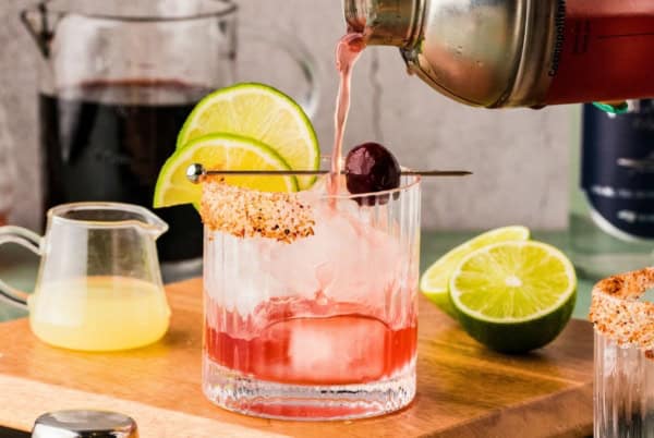 A cherry margarita is being poured from a shaker into a glass with ice, a spiced rim, and garnish of lime slices and a cherry.