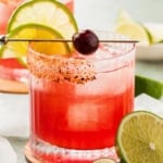 A vibrant cherry margarita drink in a glass with ice, garnished with a lime wheel, a lemon slice, a cherry, and a chili-sugar rim, with additional lime wedges nearby.