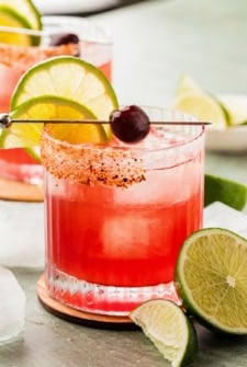 A vibrant cherry margarita drink in a glass with ice, garnished with a lime wheel, a lemon slice, a cherry, and a chili-sugar rim, with additional lime wedges nearby.