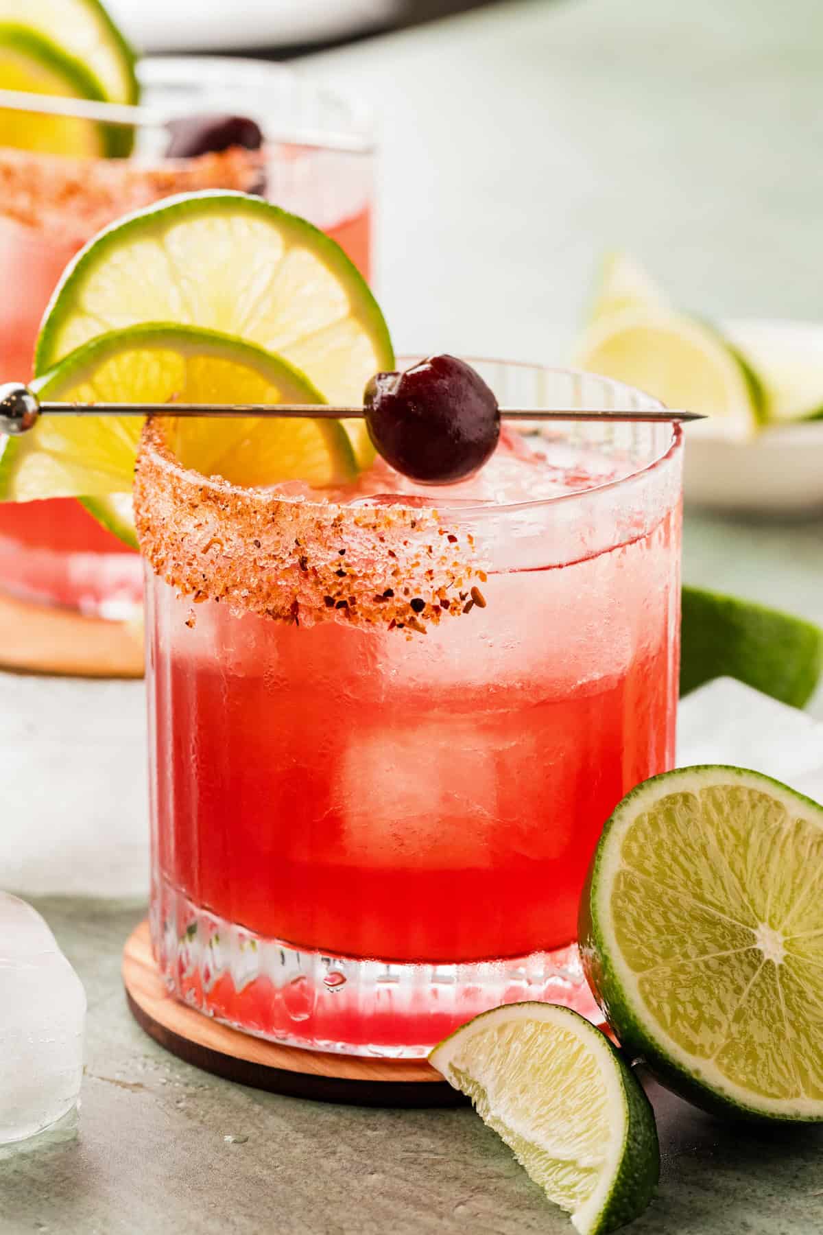Real cherry juice, no cherry liqueur, no pitting, no syrup to simmer. This Cherry Margarita takes 5 minutes and tastes like real cherries! A vibrant cherry margarita drink in a glass with ice, garnished with a lime wheel, a lemon slice, a cherry, and a chili-sugar rim, with additional lime wedges nearby.