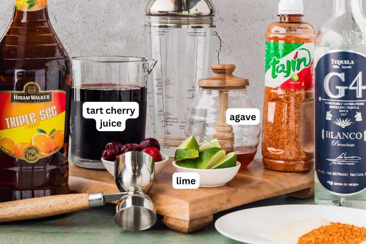 Real cherry juice, no cherry liqueur, no pitting, no syrup to simmer. This Cherry Margarita takes 5 minutes and tastes like real cherries! Ingredients for a cherry margarita recipe, including a bottle of triple sec, tart cherry juice, limes, agave, Tajin, tequila, a jigger, a shaker, and cherries, arranged on a counter with labels.