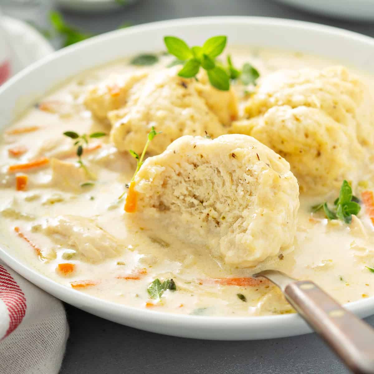 A bowl of homemade chicken and dumplings soup with a spoon lifting one of the fluffy dumplings out of the bowl of creamy soup filled with shredded chicken, carrot slices, and fresh herbs.
