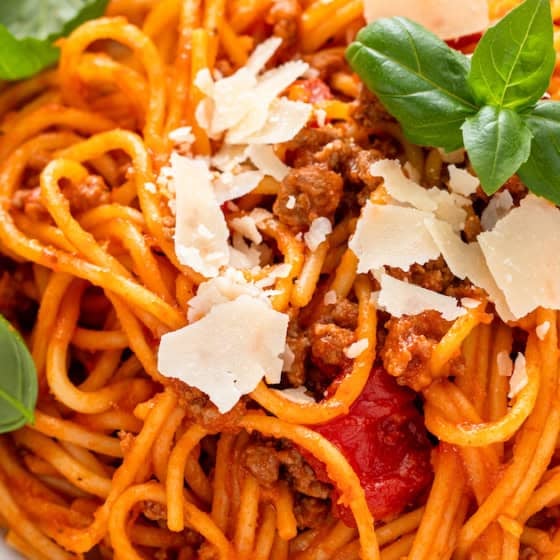 A plate of spaghetti with tomato sauce, ground meat, and cherry tomatoes, topped with shaved parmesan and fresh basil leaves.
