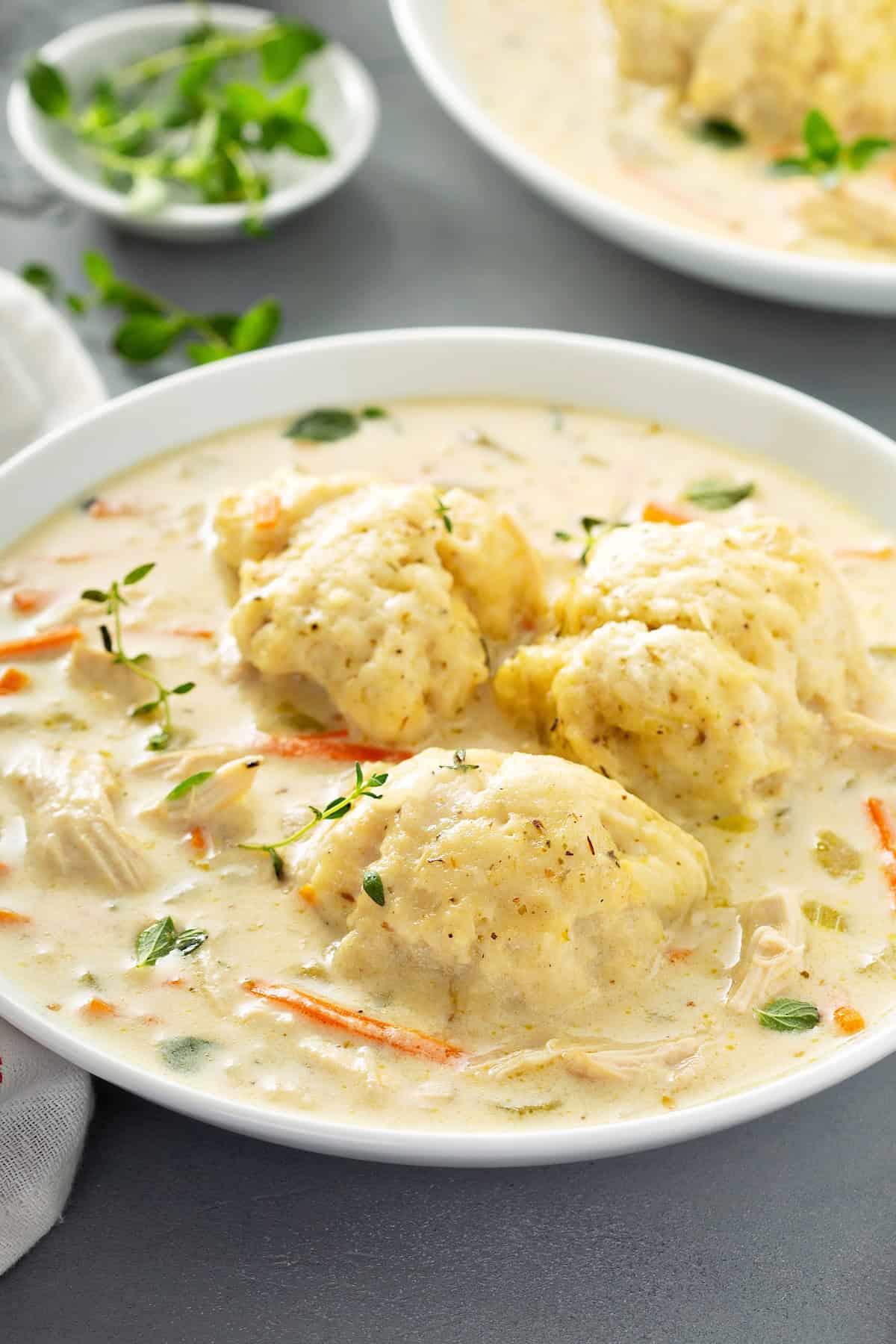 The best Chicken and Dumplings recipe you'll ever make! Ready in under an hour with a thick creamy broth, juicy chicken, and big fluffy dumplings. A white bowl filled with creamy homemade chicken and dumplings soup, topped with fluffy dumplings and garnished with fresh herbs. Vegetables and chunks of chicken are visible in the creamy broth.