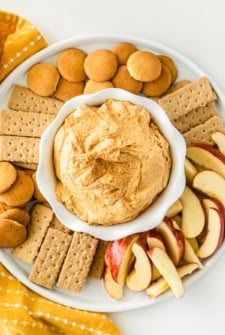 A plate with sweet potato pie dip in a bowl at the center, surrounded by vanilla wafers, graham crackers, and sliced apples, arranged neatly for dipping.