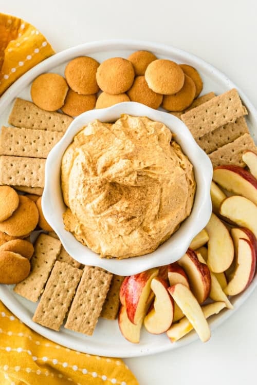 A plate with sweet potato pie dip in a bowl at the center, surrounded by vanilla wafers, graham crackers, and sliced apples, arranged neatly for dipping.