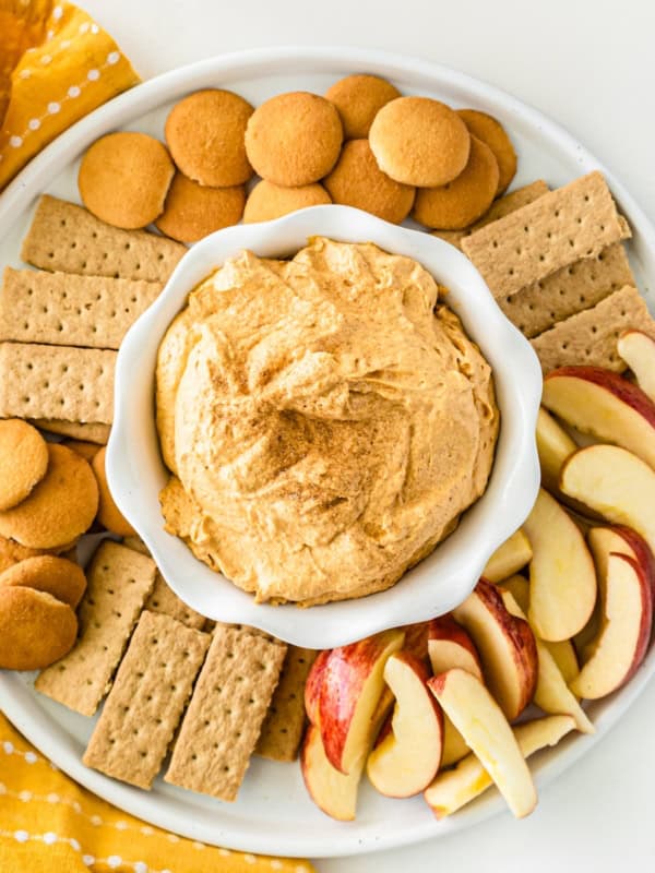 A plate with sweet potato pie dip in a bowl at the center, surrounded by vanilla wafers, graham crackers, and sliced apples, arranged neatly for dipping.