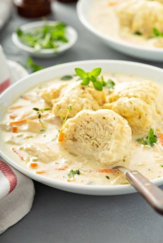 A bowl of homemade chicken and dumplings soup, topped with herbs. A fork rests on the bowl, with a dumpling partially cut open to show its fluffy inside. Carrot slices and chunks of chicken are visible in the creamy broth.