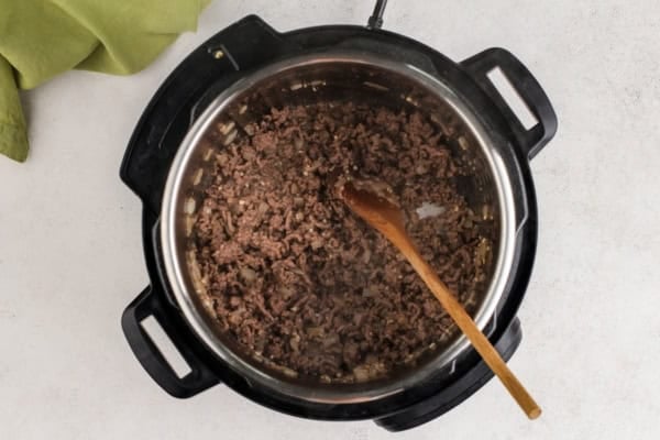 This hearty Instant Pot Spaghetti tastes like it simmered all afternoon, but it's ready in just 30 minutes and made in one single pot. Ground beef is being cooked in an Instant Pot, stirred with a wooden spoon. The pot is on a light countertop with a green cloth partially visible in the corner.